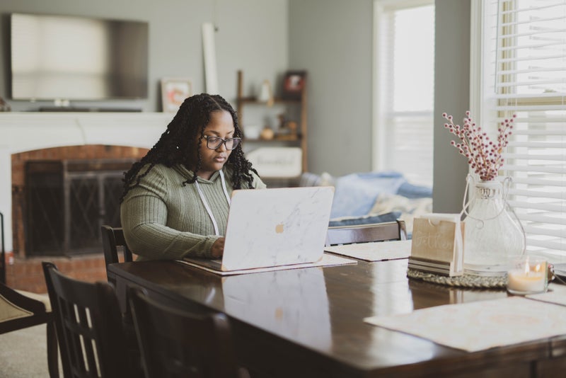 A Black woman with long braids and glasses works on a laptop at her dining room table at home.