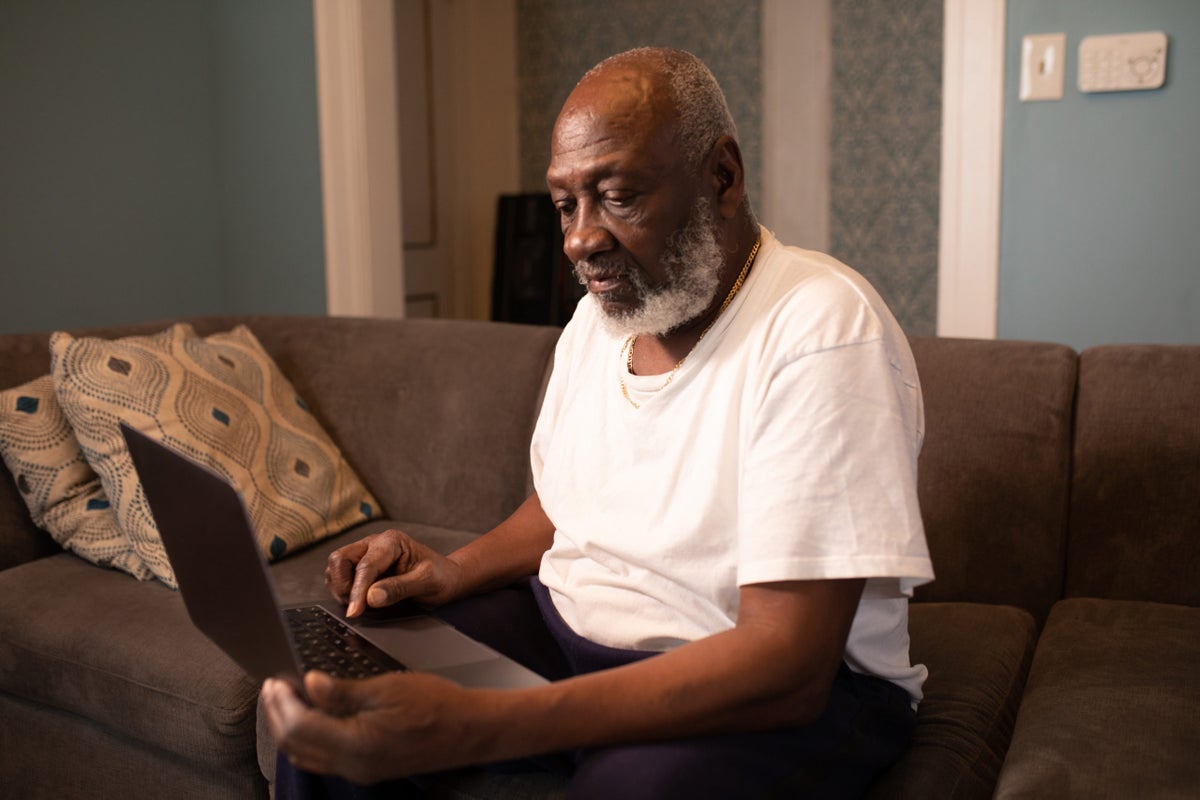 An older Black man with a white beard, in a white T-shirt and a gold chain, types on a laptop on a gray sofa at home.