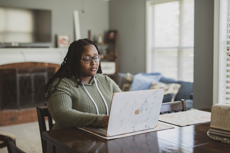 A Black woman with long twists and glasses works on a laptop at a wooden dining table in a bright living room.