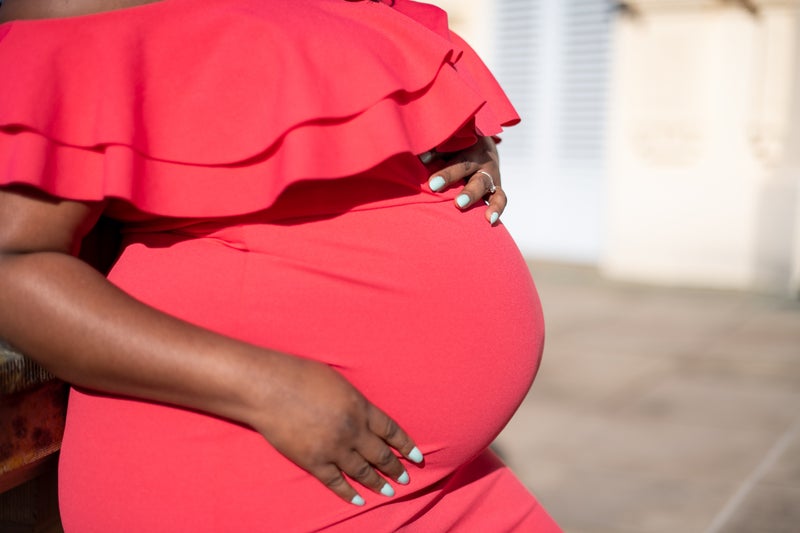 A pregnant Black woman in a red dress rests both hands on her belly.