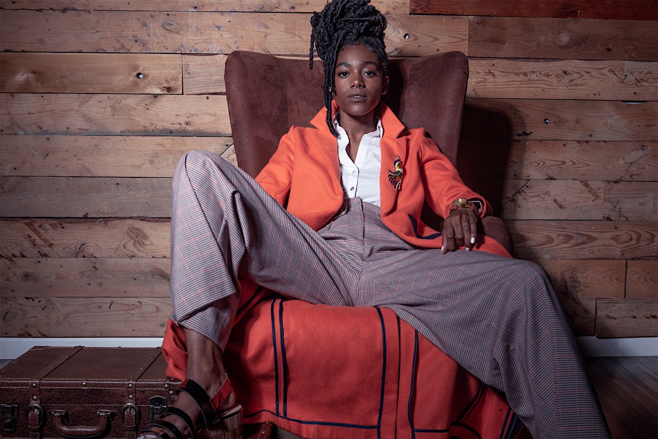 Same picture of black model wearing menswear orange long coat and checkered suit pants.